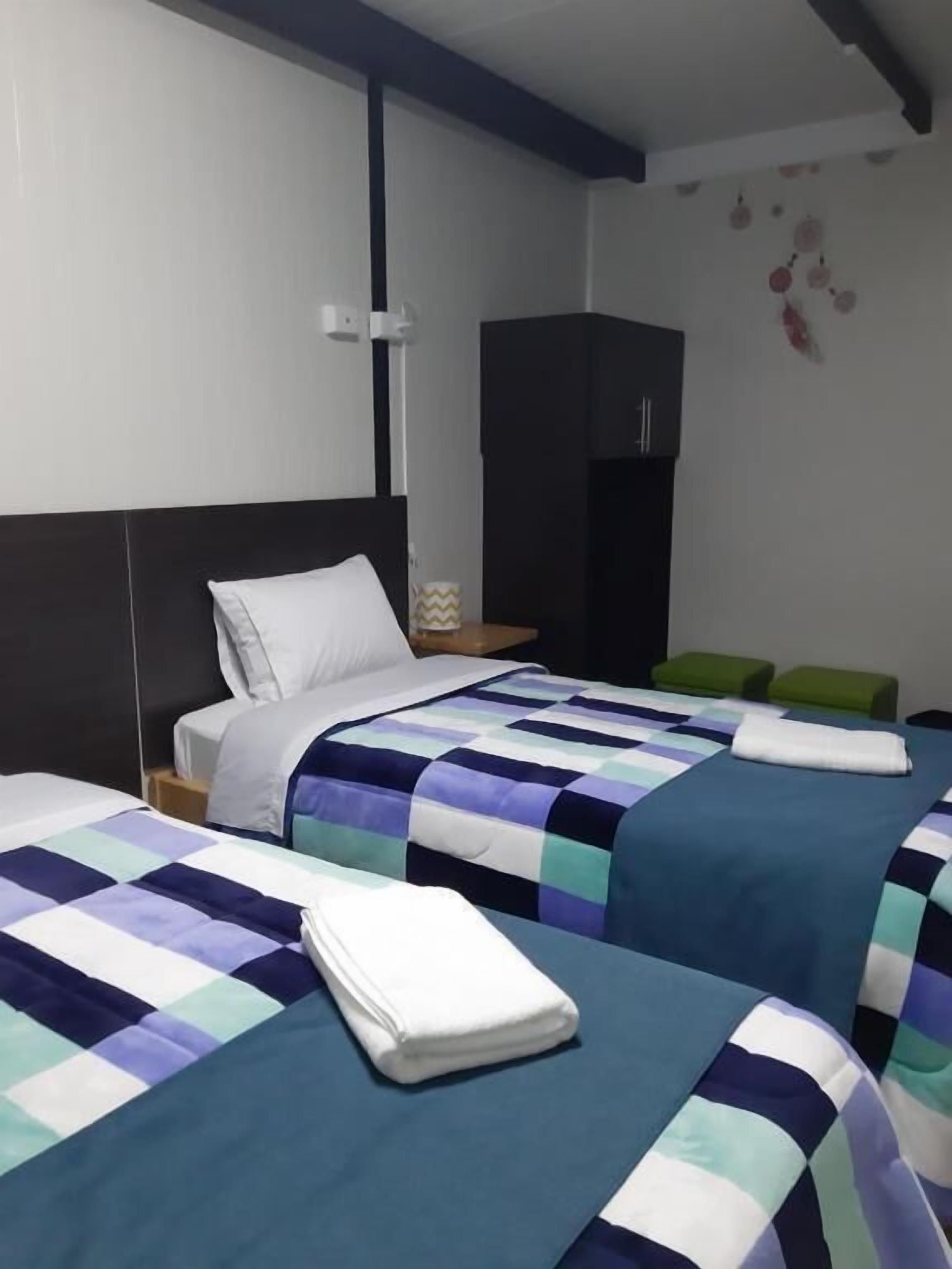 A hotel room with two beds, each with a blue and white checkered bedspread, a white pillow, and a neatly folded white towel on the bed. There is a green bench in the corner and a black wardrobe in the background.