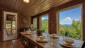 Una mesa de comedor de madera dispuesta con platos y vasos, un reloj en la pared y vistas a la vegetación exterior.