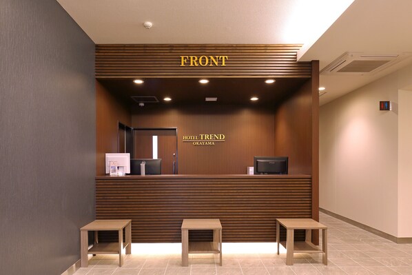 Un espace d’accueil avec un comptoir en bois, une enseigne indiquant « ENTRÉE » et une autre indiquant « HÔTEL TREND OKAYAMA ». Trois bancs en bois sont placés devant le comptoir.