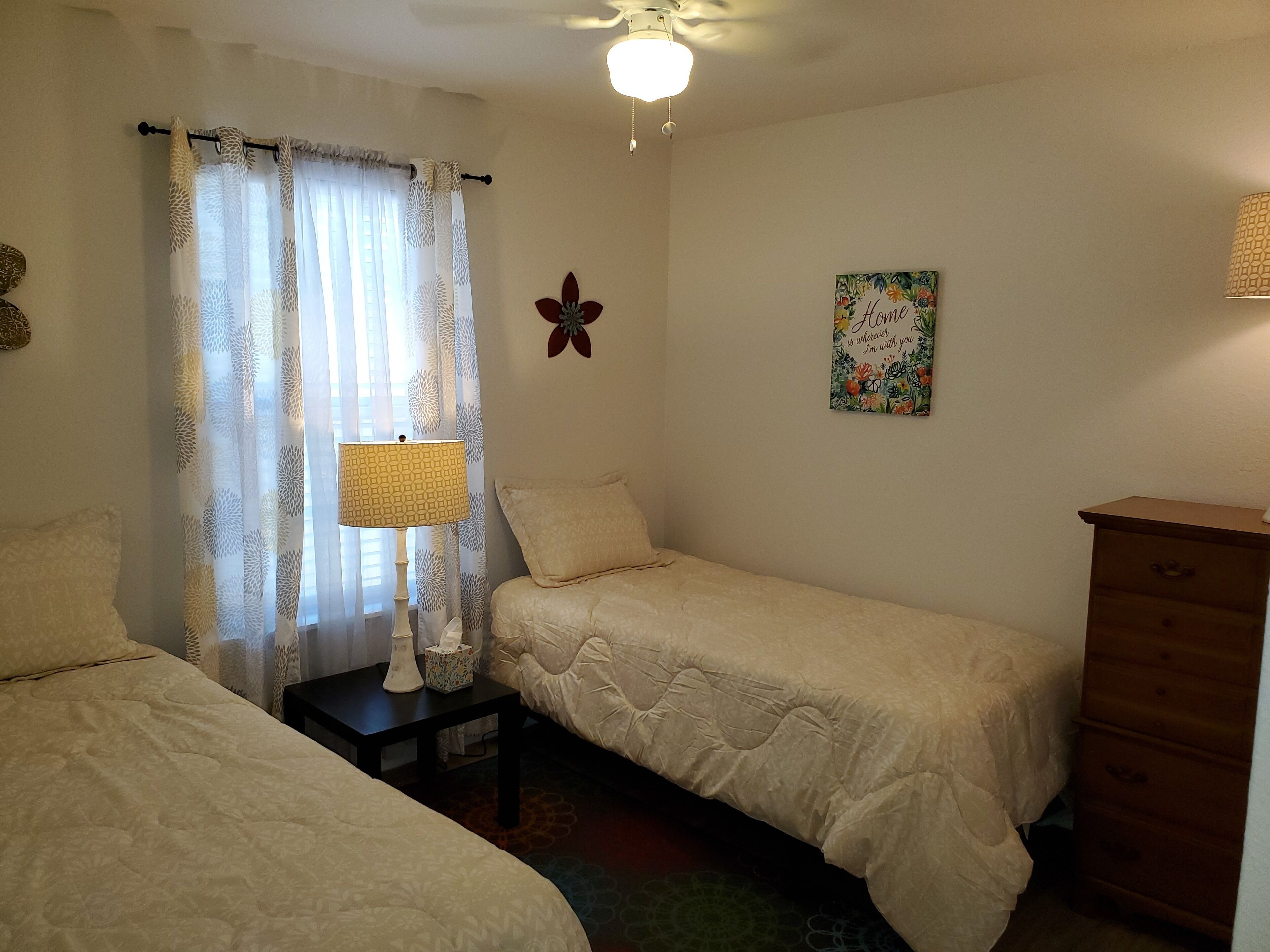 A bedroom with two single beds, a bedside table with a lamp, a dresser, a decorative butterfly, and a 'Home is where you are with me' sign.