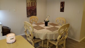 Un coin repas avec une table dressée pour quatre personnes, entourée de chaises en bois aux motifs complexes dans le dossier. Une nappe blanche recouvre la table, et une seule orchidée est placée dans un vase noir posé sur la table. Un luminaire suspendu se trouve au-dessus de la table, et un tableau encadré est accroché au mur derrière.