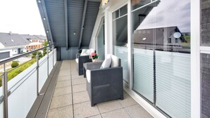 Een balkon met buitenmeubilair, uitzicht op omliggende huizen, en een overdekt gedeelte met een schuin dak.