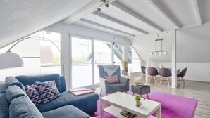 Een modern loft appartement met een hoge plafond, grote ramen, een gezellige zithoek met een bank en fauteuils, een eettafel, en een trap naar een hoger niveau.