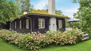 Una casa de madera negra con una chimenea blanca, rodeada de abundante follaje y flores rosadas.