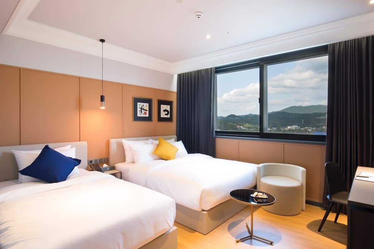 Ein Hotelzimmer mit zwei Betten, einem kleinen Tisch, einem Sessel und Blick auf die Stadt durch große Fenster.