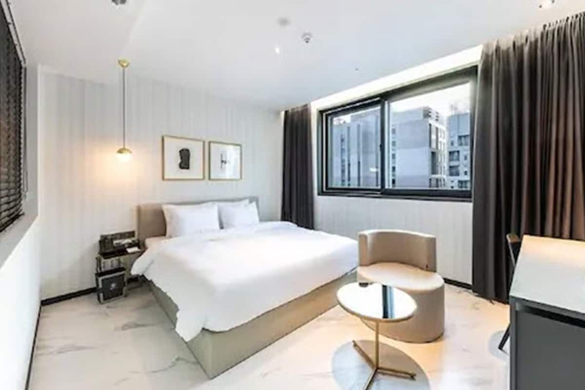 Ein modernes Hotelzimmer mit einem großen Bett, einer runden Sitzecke, einem Schreibtisch und einem Fenster mit Blick auf die Stadt.