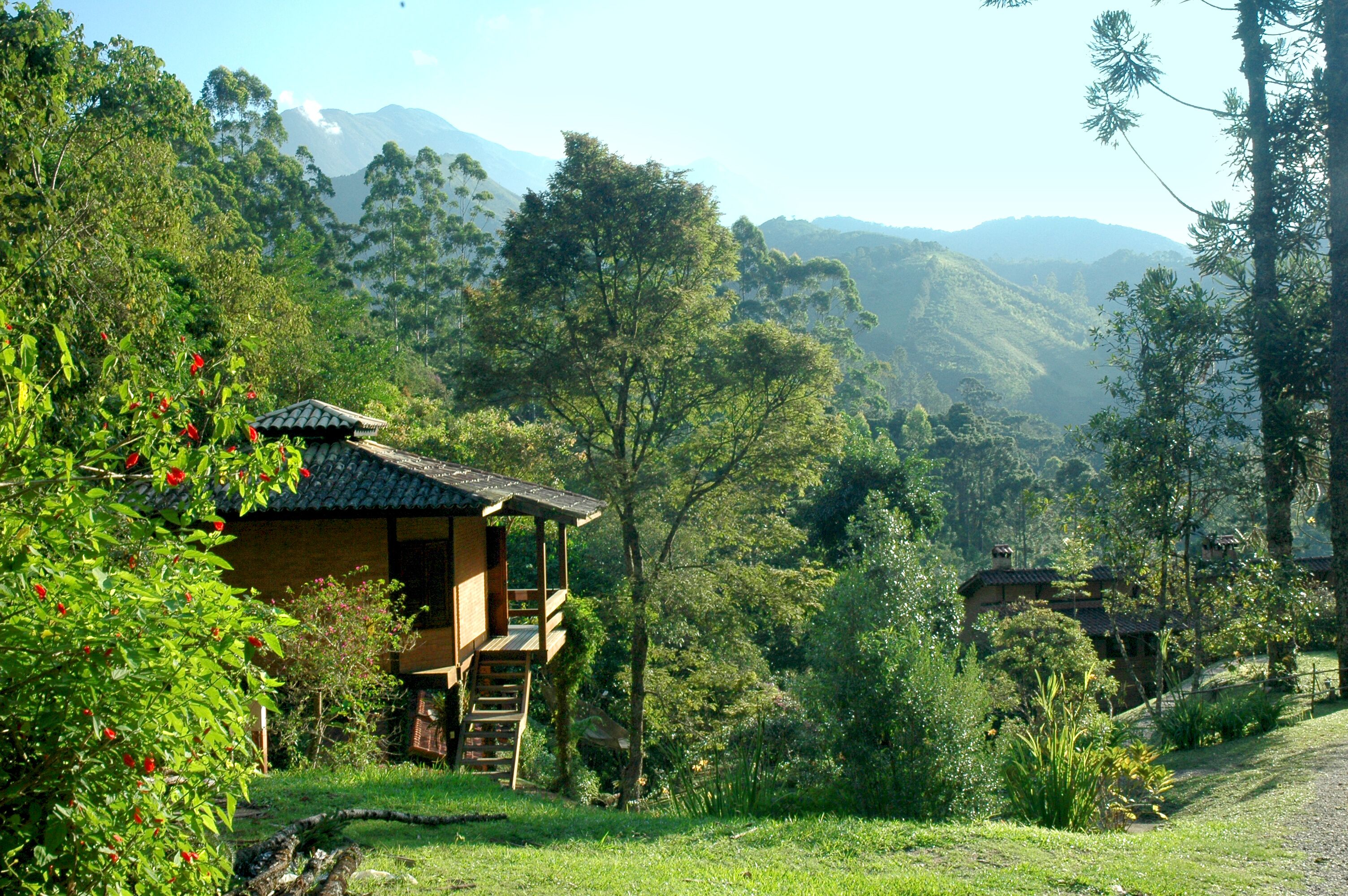 A traditional wooden house with a balcony, surrounded by lush greenery and mountains in the background.