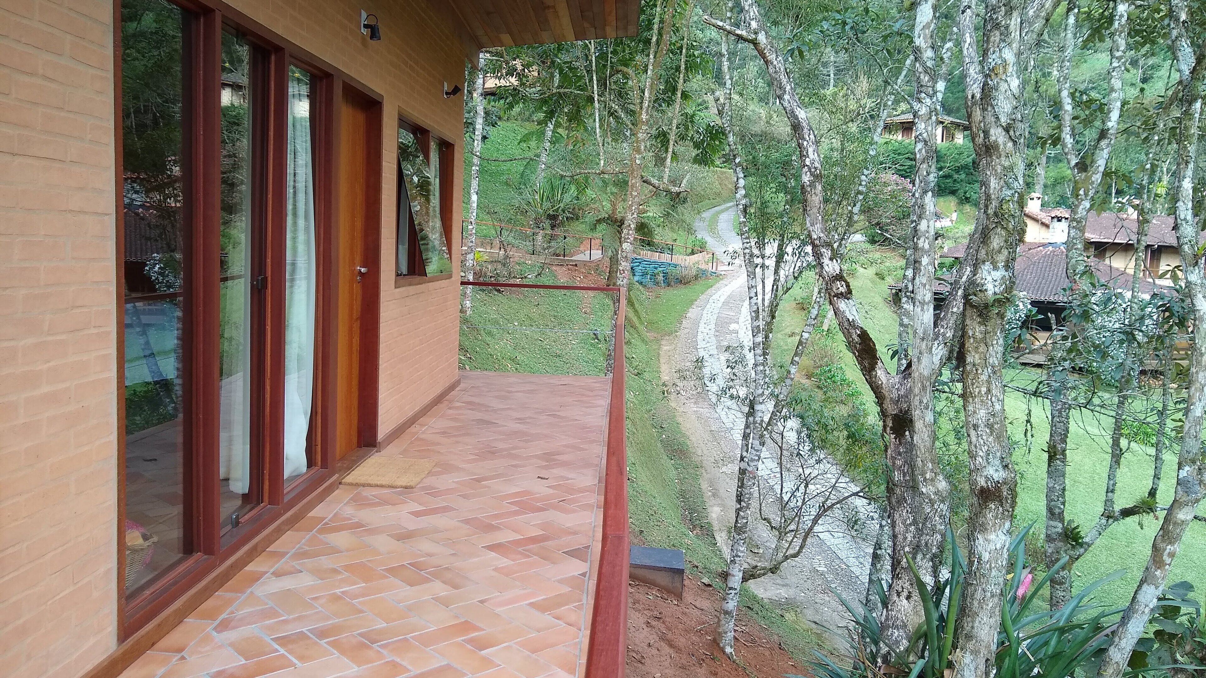 A covered outdoor area with a brick floor, large glass doors, and a view of a green landscape with trees and a path.