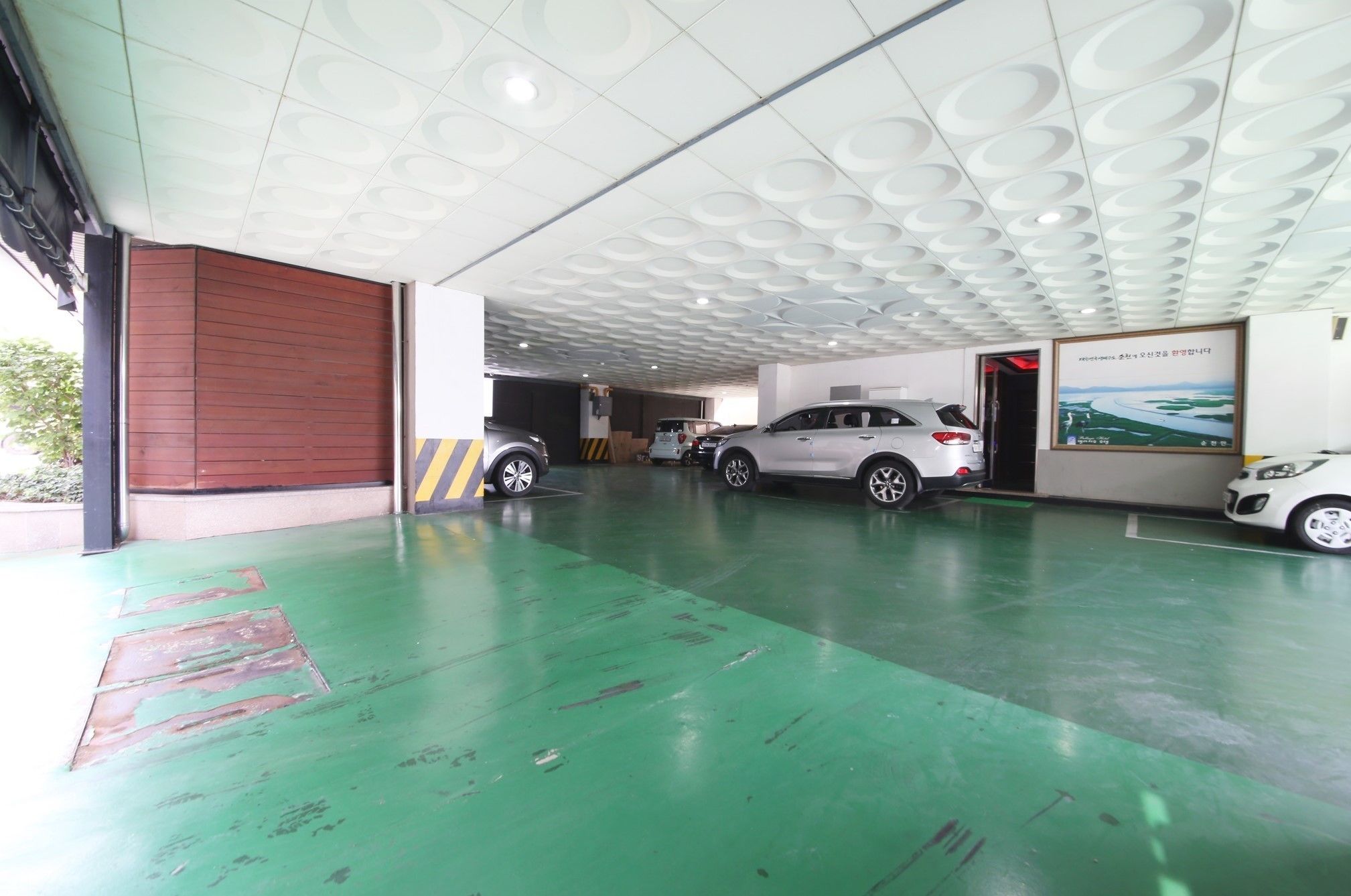 A parking garage with a green floor, white ceiling with circular patterns, and a large wooden door on the left.