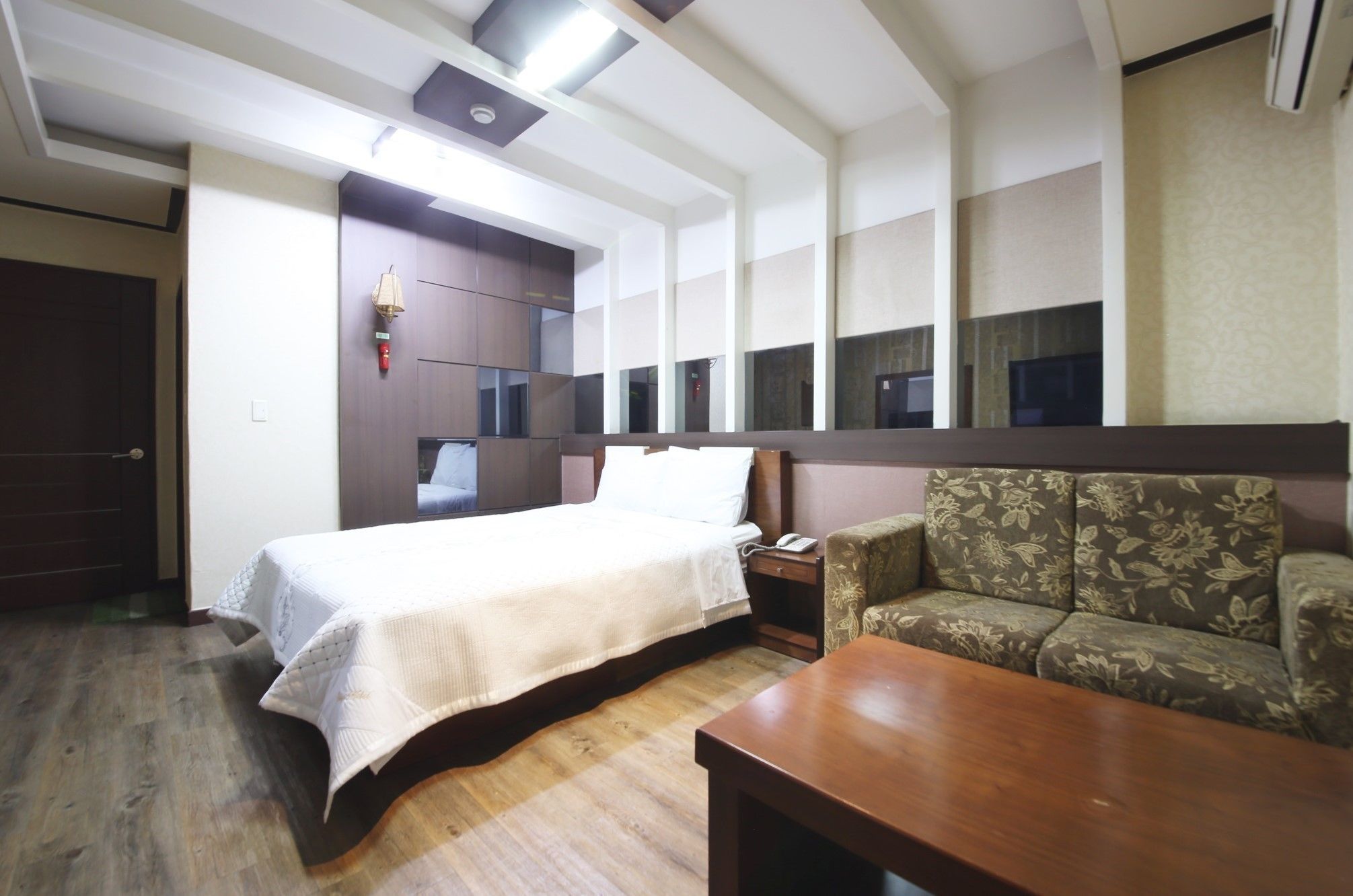 A hotel room with a bed, a sofa, a wooden coffee table, a nightstand, and a wall-mounted TV.