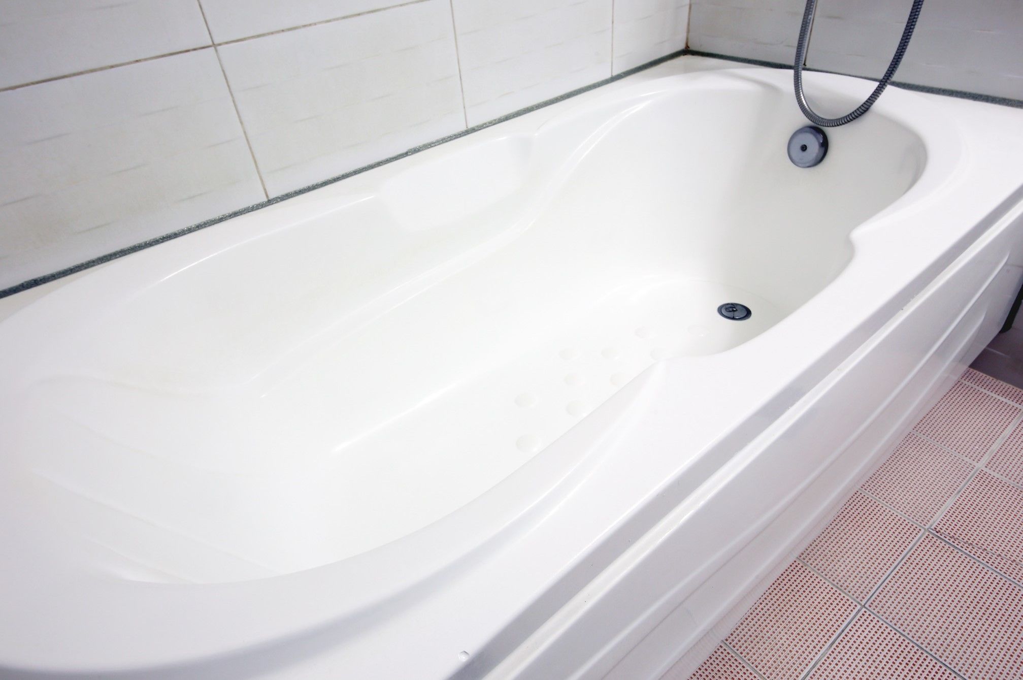 Eine weiße Badewanne mit Ablass, Duschschlauch und gefliestem Boden.