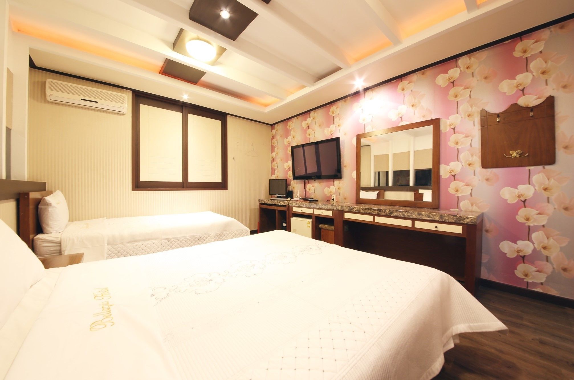 A hotel room with two beds, a TV, a mirror, and a vanity with a floral wallpaper.