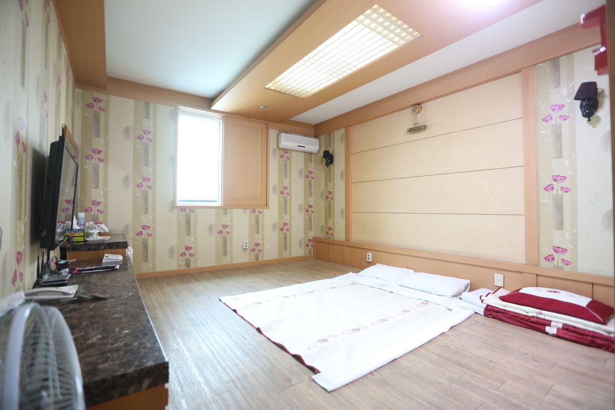 A bedroom with a large bed, a desk with a fan, a window, and a ceiling light.