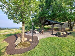 Un'area salotto esterna coperta, con tavolo e sedie, circondata da un giardino curato con rocce e un albero.