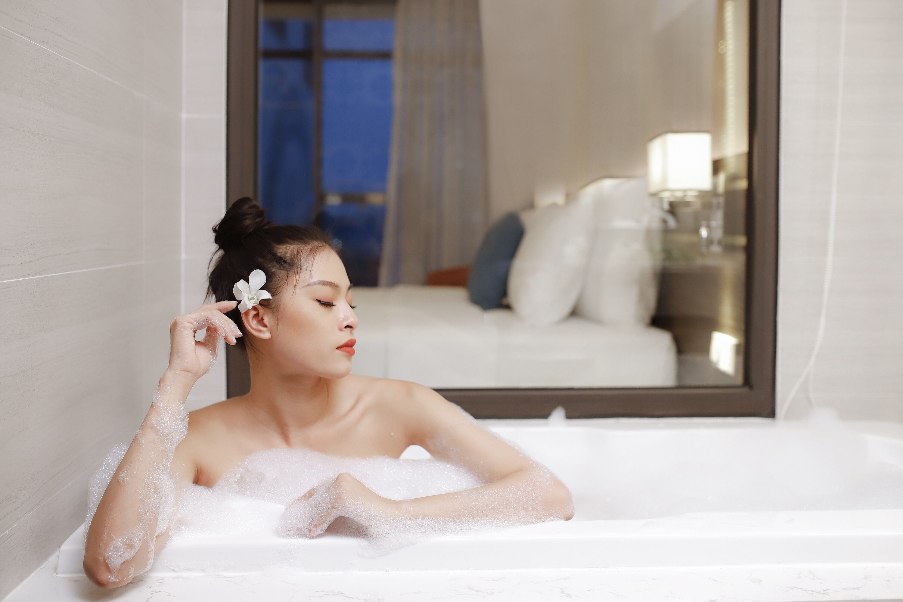 A person in a bathtub with a mirror reflecting a bedroom.