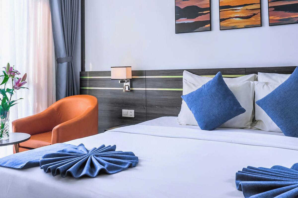 A hotel room with a bed, a chair, a nightstand, and a lamp. There are three paintings on the wall.