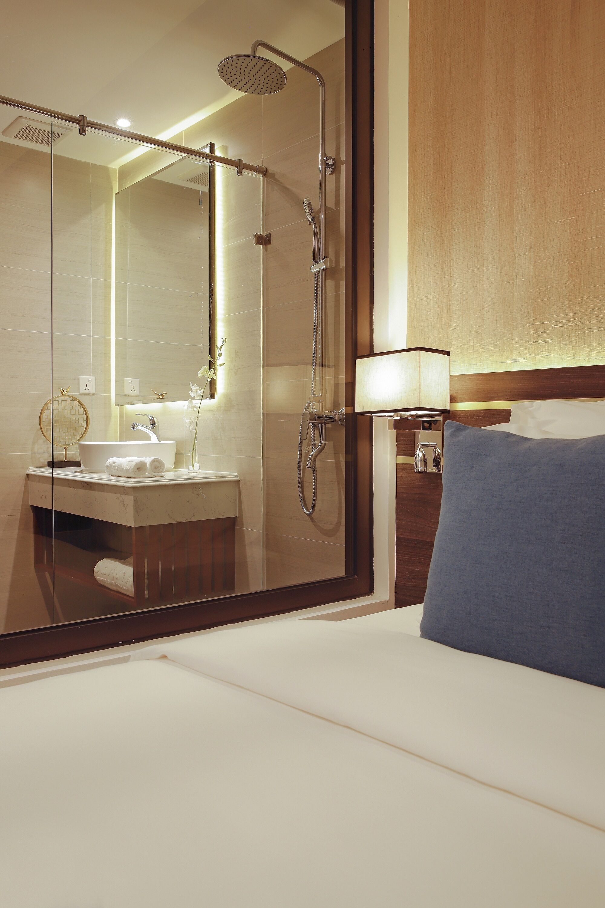 A hotel room with a glass-enclosed shower, a sink with a mirror, a bedside lamp, and a blue pillow on the bed.