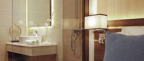 A hotel room with a glass-enclosed shower, a sink with a mirror, a bedside lamp, and a blue pillow on the bed.