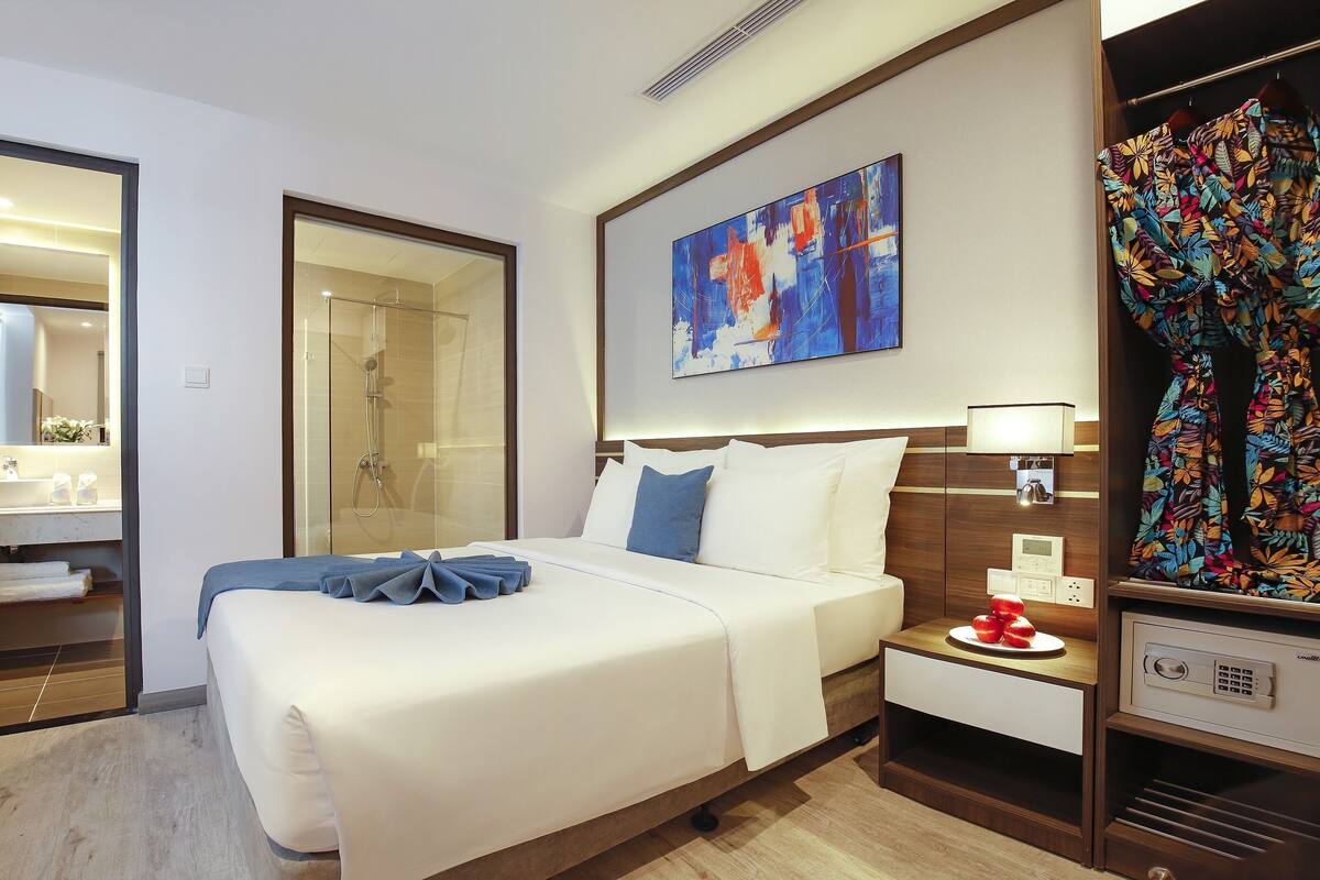 A hotel room with a large bed, a bedside table with a lamp, a shower, a bathroom with a mirror, and a colorful artwork on the wall.