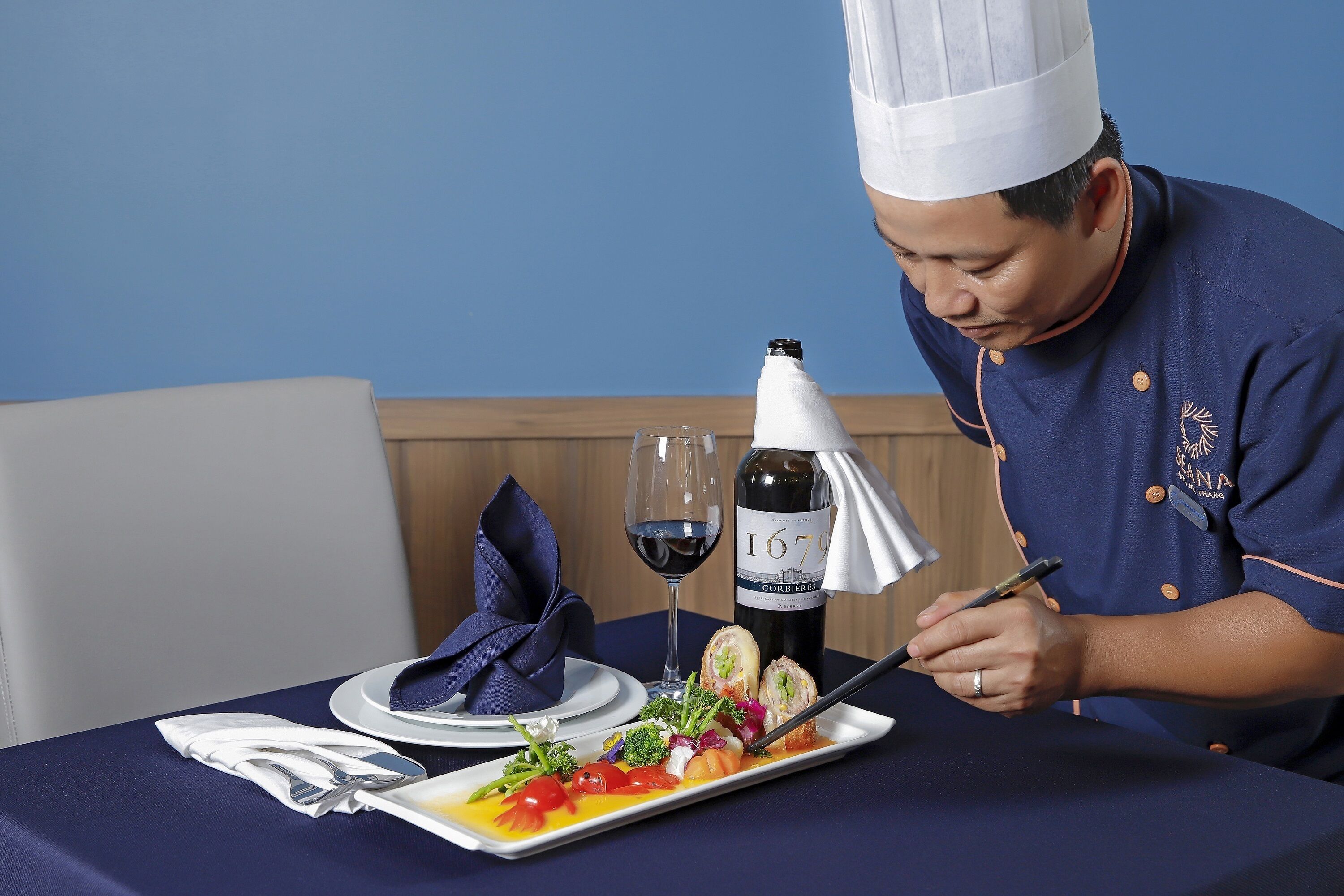 A chef in uniform preparing a dish at a table with a wine bottle and a glass.