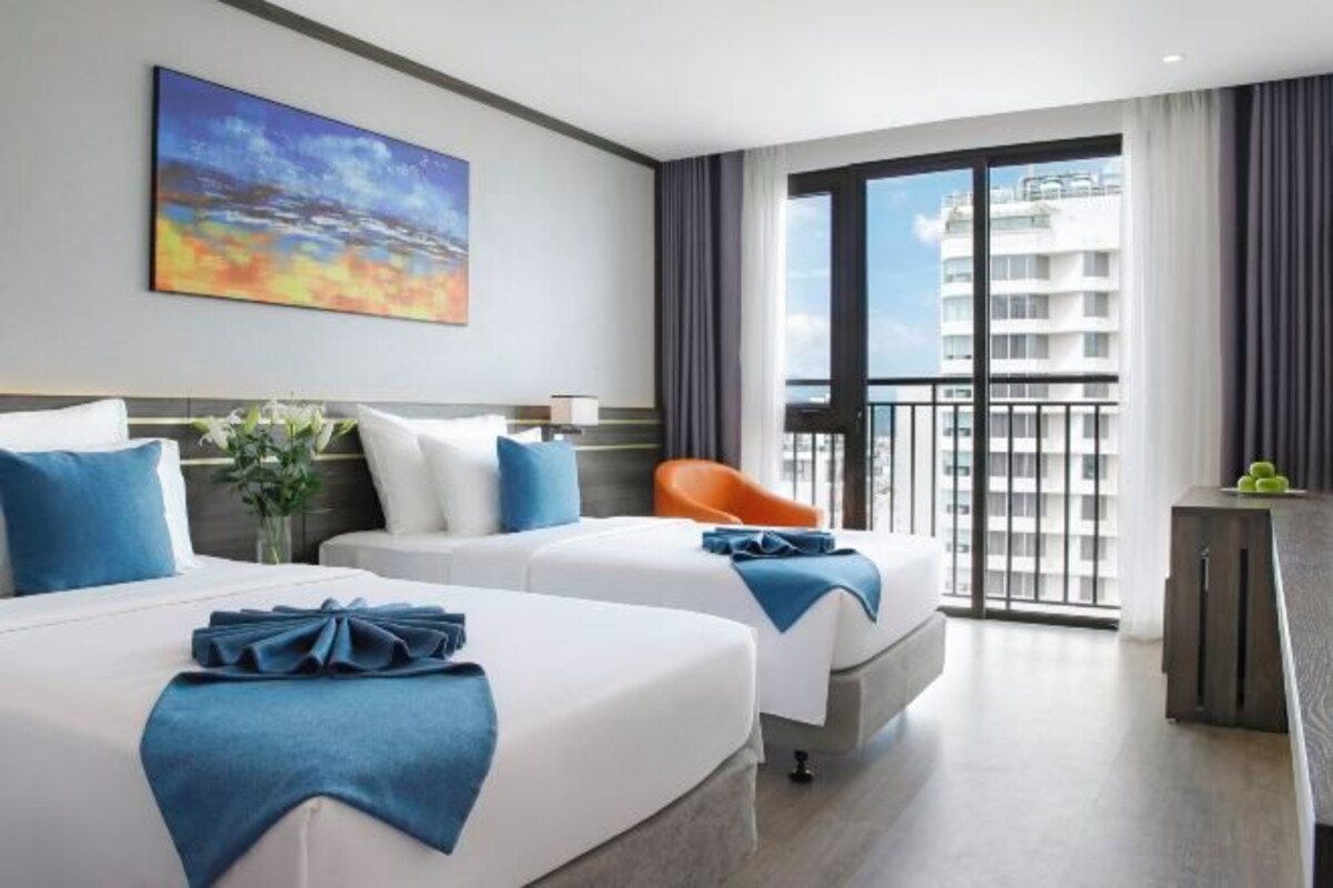 Une chambre d’hôtel avec deux lits, une grande fenêtre donnant sur un immeuble, un tableau accroché au mur et une chaise.