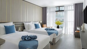 Een hotelkamer met twee bedden, een groot raam met gordijnen, een vaas met bloemen en een bureau met een lamp.