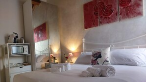 Ein ordentlich gemachtes Bett mit weißen Leinentüchern und aufgerollten Handtüchern, ein Nachttisch mit Lampe, eine Mikrowelle auf einem Regal und ein Spiegel, der den Raum reflektiert.