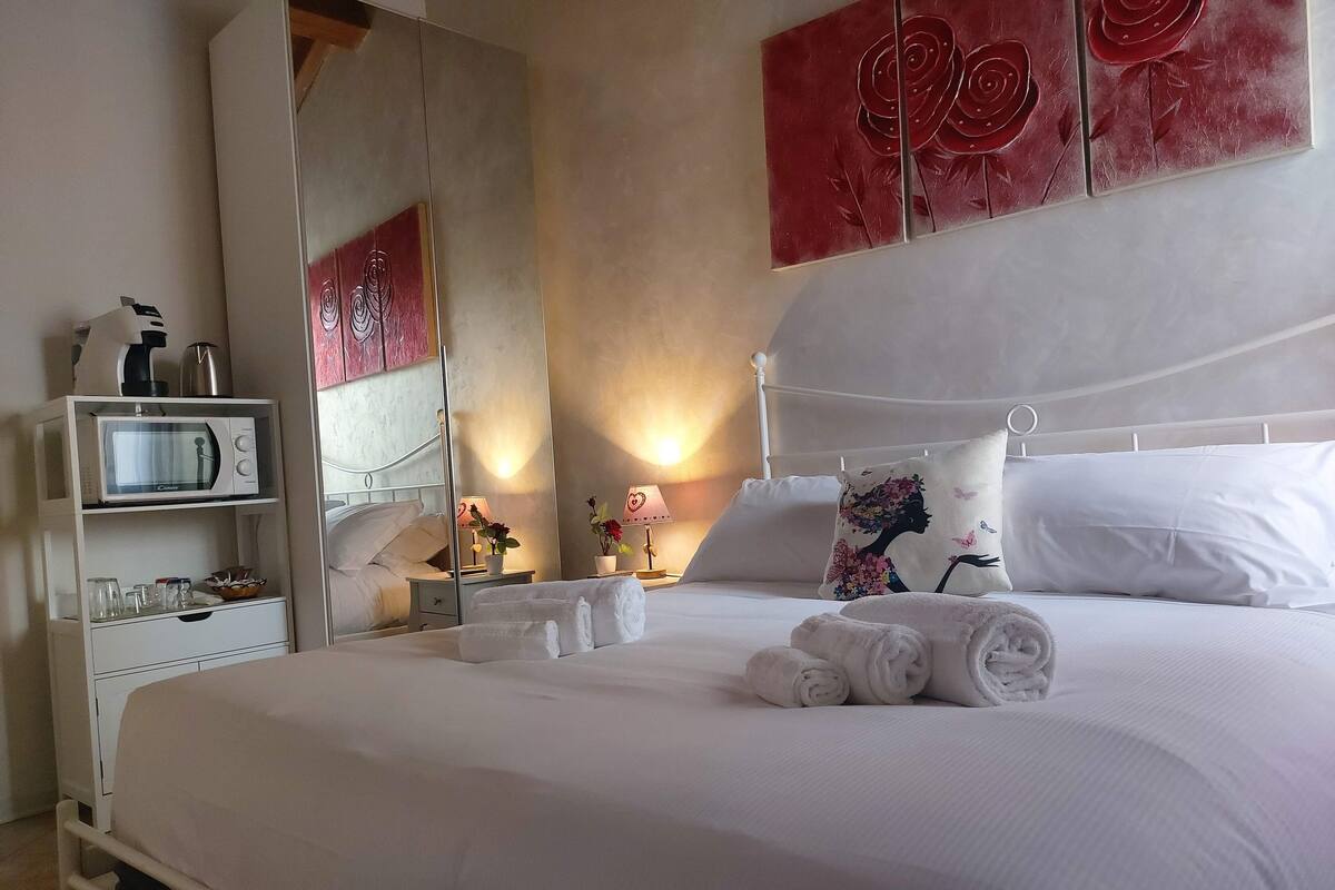 A neatly made bed with white linens and rolled towels, a bedside table with a lamp, a microwave on a shelf, and a mirror reflecting the room.