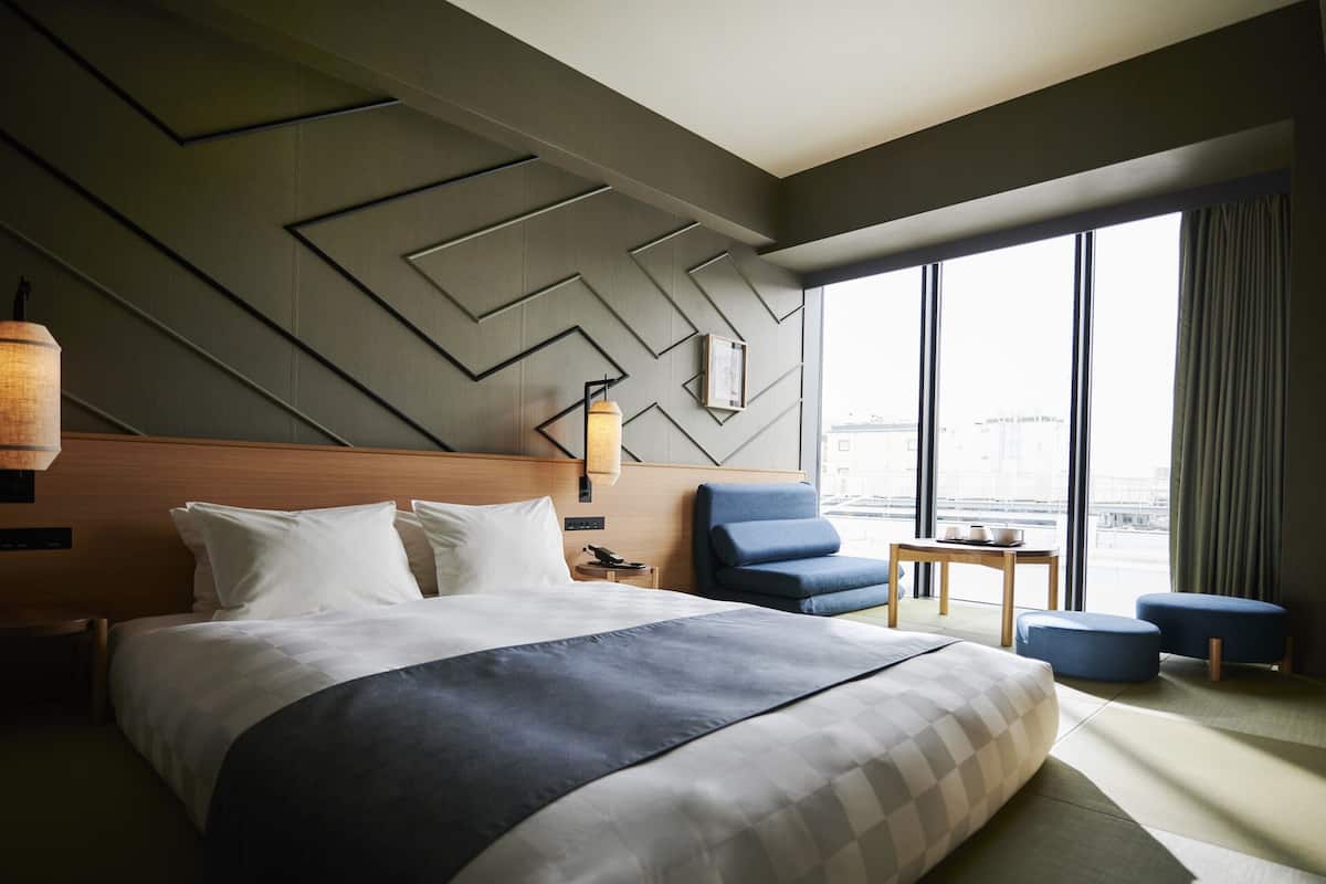 Una habitación de hotel moderna con una cama grande, una zona de estar con una mesa y vistas a la ciudad a través de amplios ventanales.