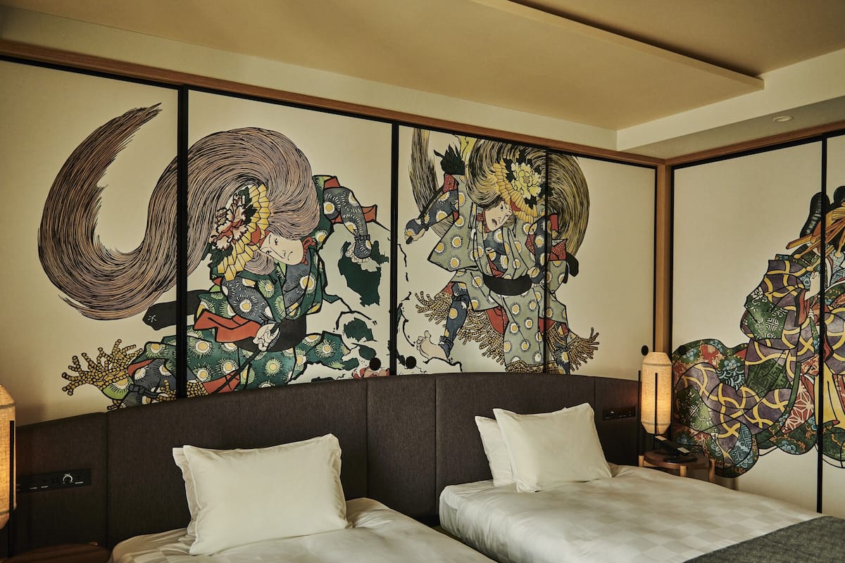 Habitación de hotel con dos camas, una obra de arte tradicional de gran tamaño en la pared y una mesita de noche de madera con una lámpara.