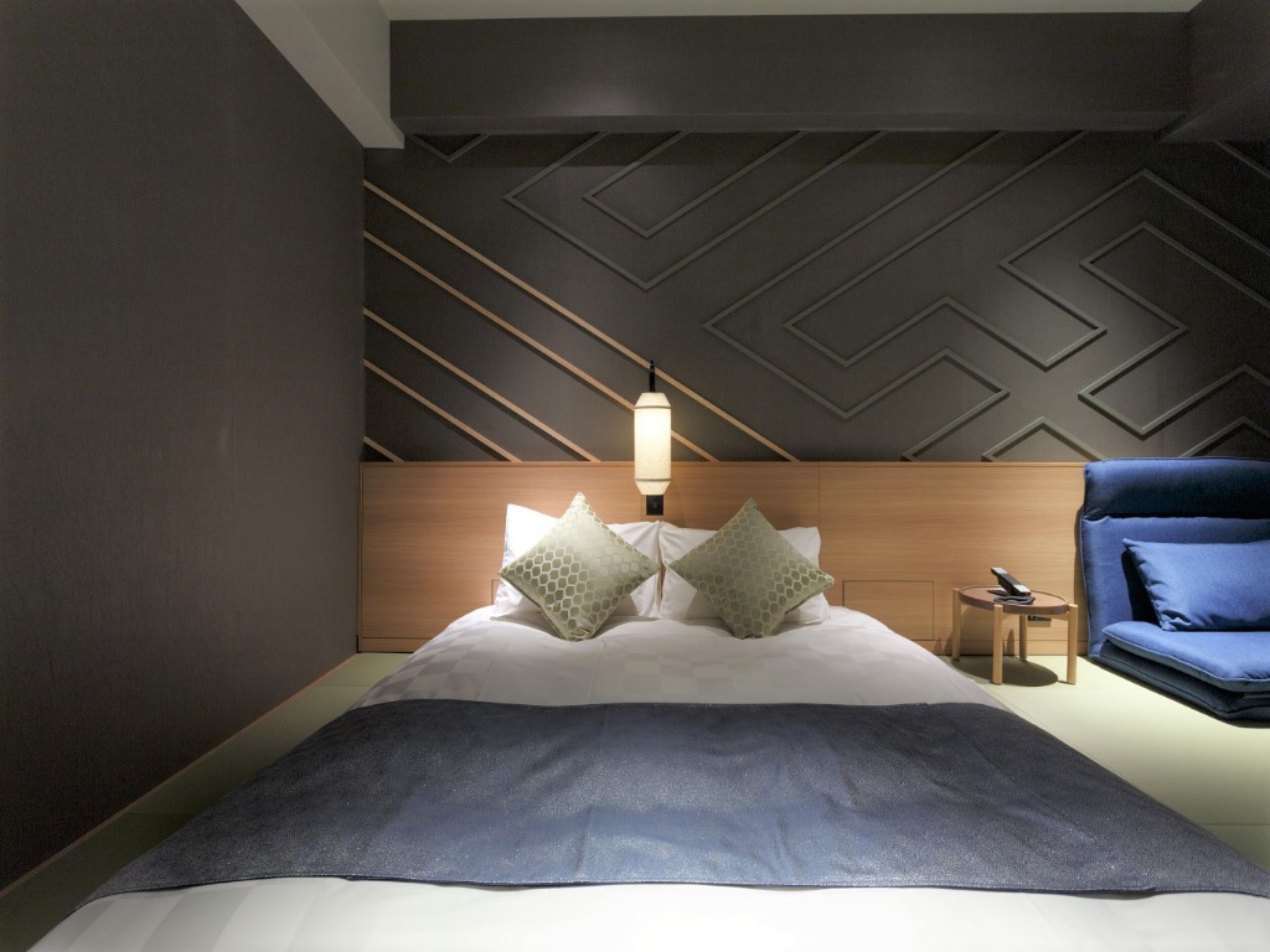 A modern hotel room with a bed, a nightstand, a lamp, and a blue armchair.