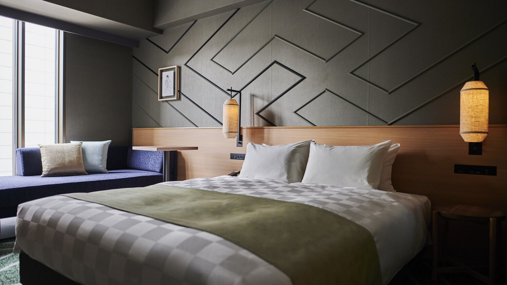 A hotel room with a bed, a sofa, a wall with geometric patterns, and a hanging light fixture.