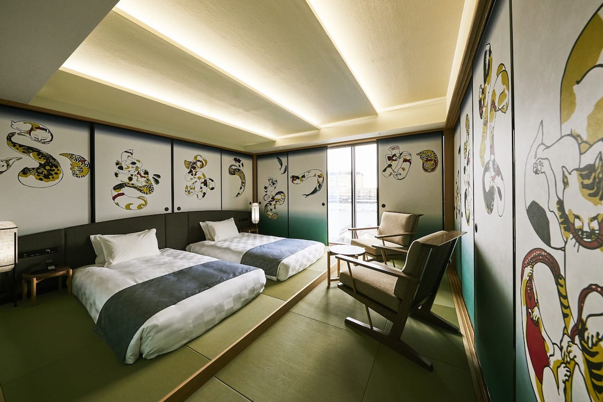 Habitación de hotel con dos camas, una silla, una mesita y obras de arte en las paredes.