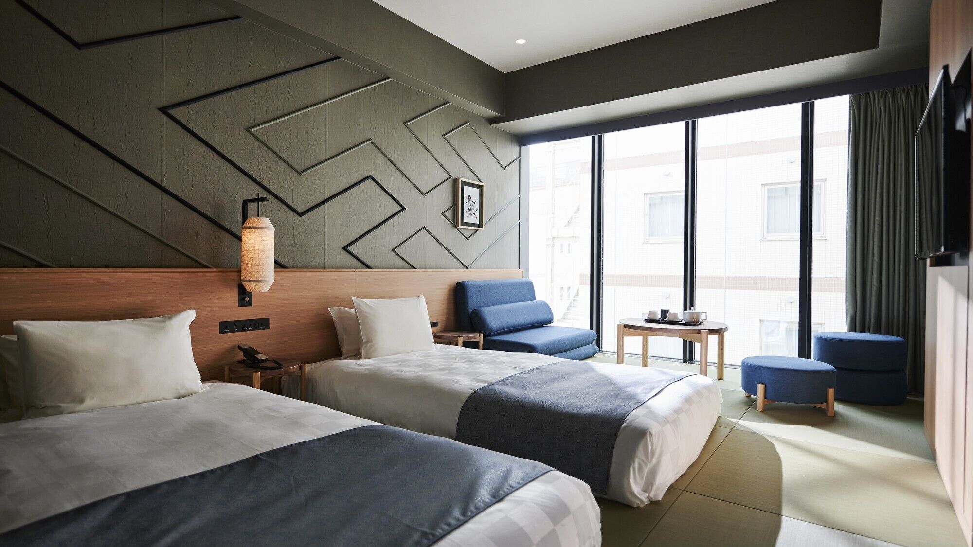 A hotel room with two beds, a small table, a chair, and a large window.