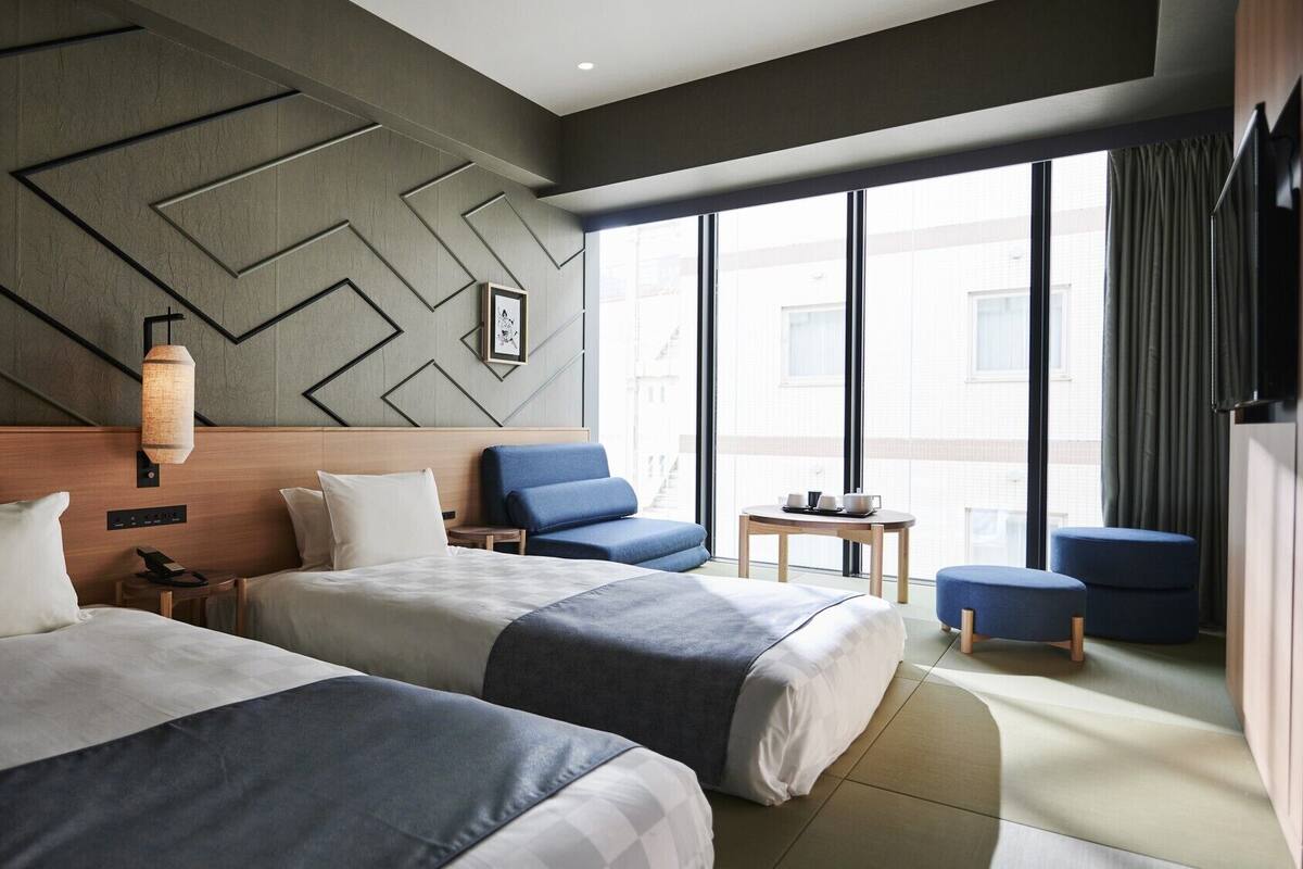 A hotel room with two beds, a small table, a chair, and a large window.