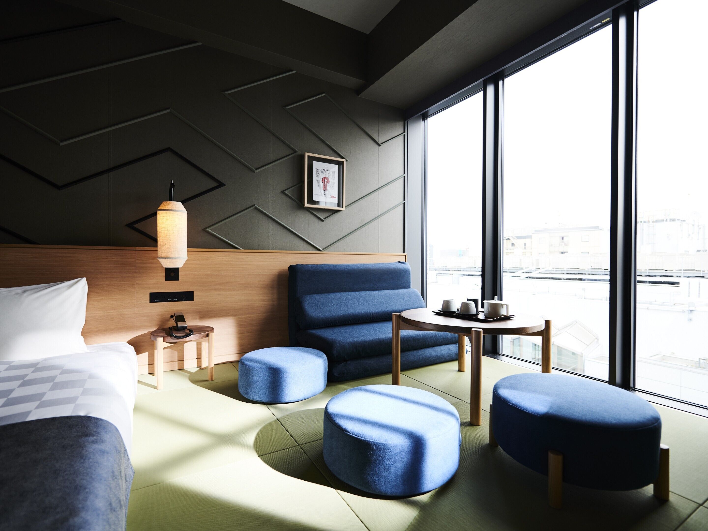 A modern hotel room with a bed, a blue armchair, a small table with cups, and a view of the city through large windows.