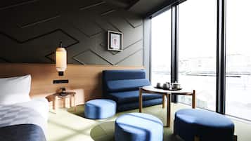 A modern hotel room with a bed, a blue armchair, a small table with cups, and a view of the city through large windows.