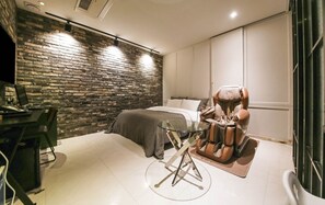 A modern bedroom with a brick wall, a bed, a massage chair, and a desk with a computer. - Lexy Hotel (Daejeon)