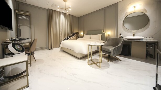 A modern hotel room with a large bed, a round mirror, a bedside table, a chair, a desk with a computer, and a bathroom with a sink and mirror. - Lexy Hotel (Daejeon)