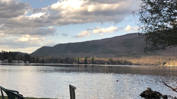 A lakeside view with a fire pit and chairs, surrounded by mountains and trees.