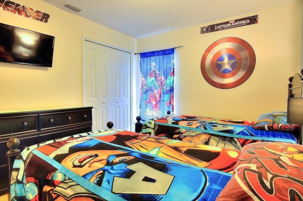 Een slaapkamer met een bed, voorzien van een dekbedovertrek met superheldenprint, een wandgemonteerde flatscreen-tv en een Captain America schild aan de muur.