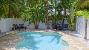 Una piscina con borde curvo, rodeada por un solárium empedrado. Hay dos sillas de metal negro y una mesa con mantel estampado.