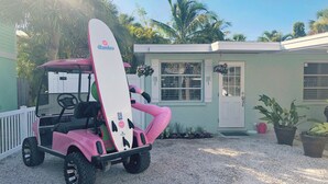 Un carrito de golf rosa con una tabla de surf sujeta, estacionado frente a una casa con puerta y ventanas, rodeado de plantas en macetas y palmeras.