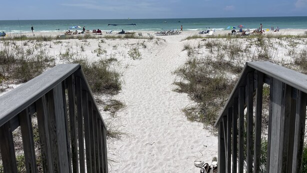 A sandy beach with a wooden walkway leading to the water. Beach chairs and umbrellas are visible. - Spacious 3 Bedroom, 2 1/2 bath Condo, Steps to the beach! 2 pools and Hot Tub! (Bradenton Beach)