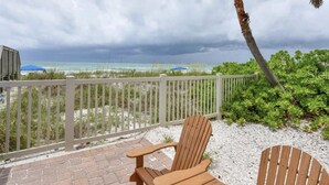A wooden deck with two Adirondack chairs overlooking a beach with a white fence and palm trees. - Spacious 3 Bedroom, 2 1/2 bath Condo, Steps to the beach! 2 pools and Hot Tub! (Bradenton Beach)