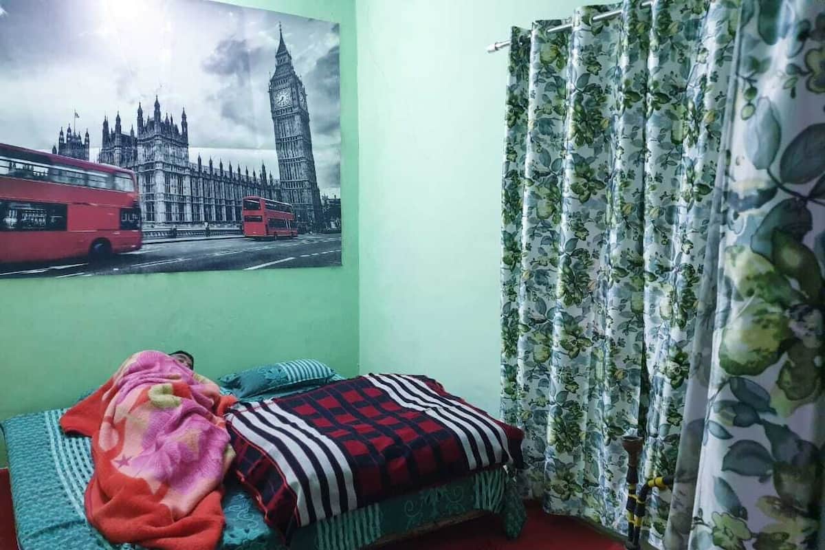 Ein Zimmer mit einem Bett, einer roten Decke, einem gemusterten Überwurf, einer Wand mit einem Bild eines Gebäudes und floralen Vorhängen.