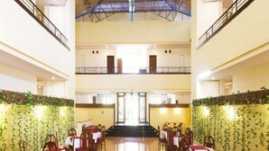 A spacious indoor area with multiple dining tables and chairs, a high ceiling with a metal framework, and a central staircase leading to an upper level. - The Dhanhills - A Valley View Hotel In Panchgani (Mahabaleshwar)