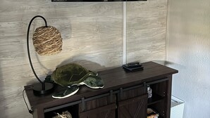 A modern living room with a dark wood entertainment center, a flat-screen TV mounted above it, and a woven wall hanging. - Reel’em Inn - a Waterfront Home on the Canal (Aransas Pass)