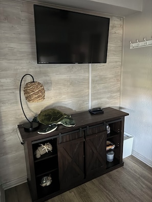 A modern living room with a dark wood entertainment center, a flat-screen TV mounted above it, and a woven wall hanging. - Reel’em Inn - a Waterfront Home on the Canal (Aransas Pass)