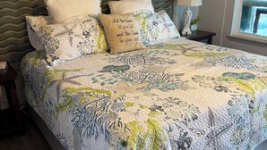 A neatly made bed with floral bedding, a headboard with a wavy design, and a bedside table with a lamp and a plant. - Reel’em Inn - a Waterfront Home on the Canal (Aransas Pass)